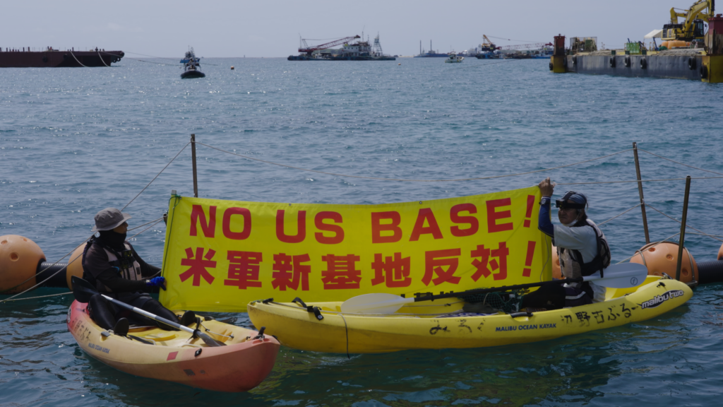 Demonstranten in kajaks met een spandoek ‘NO US BASE!’ in Okinawa, uit Earth’s Greatest Enemy.